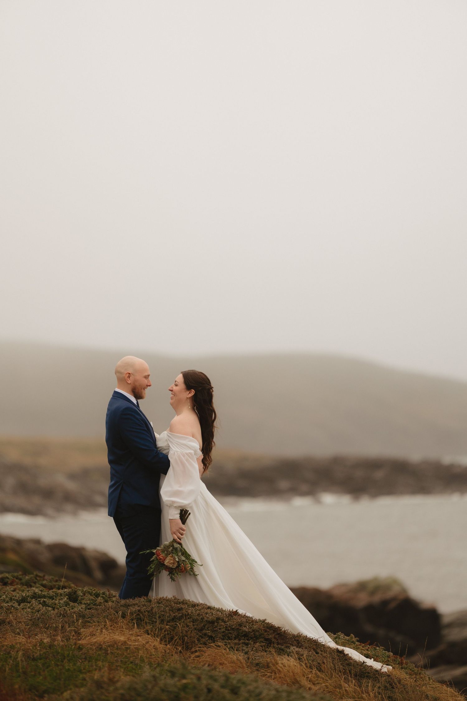 A couple in wedding attire embraces on a misty coastal cliff overlooking the ocean on their special day.