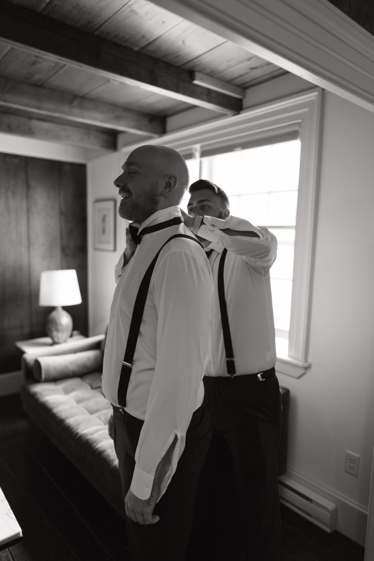 Two people in white dress shirts and dark suspenders stand in conversation near a window in a bright room.