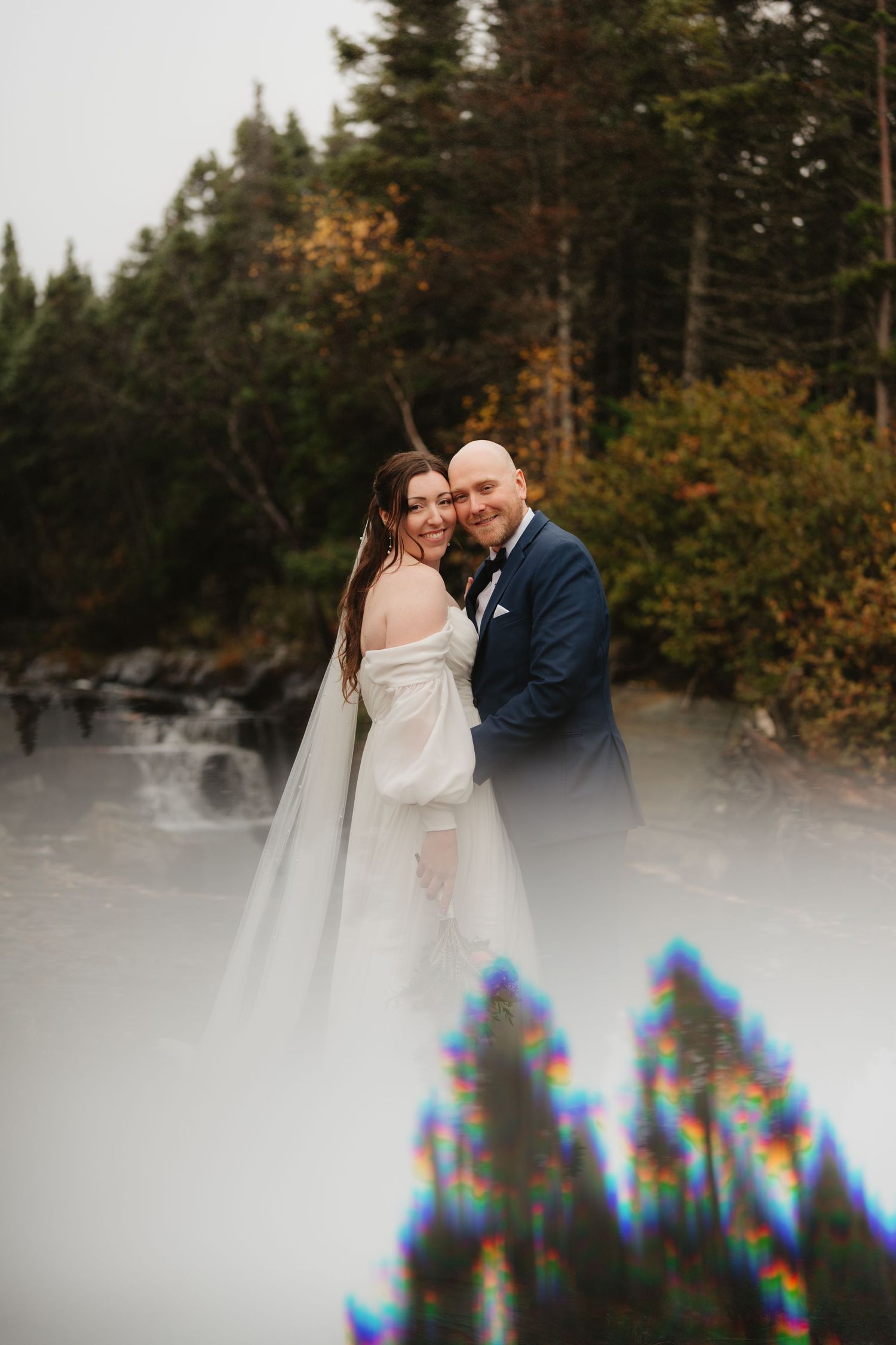 A couple shares an intimate moment in a snowy forest setting with evergreen trees and autumn foliage in the background.