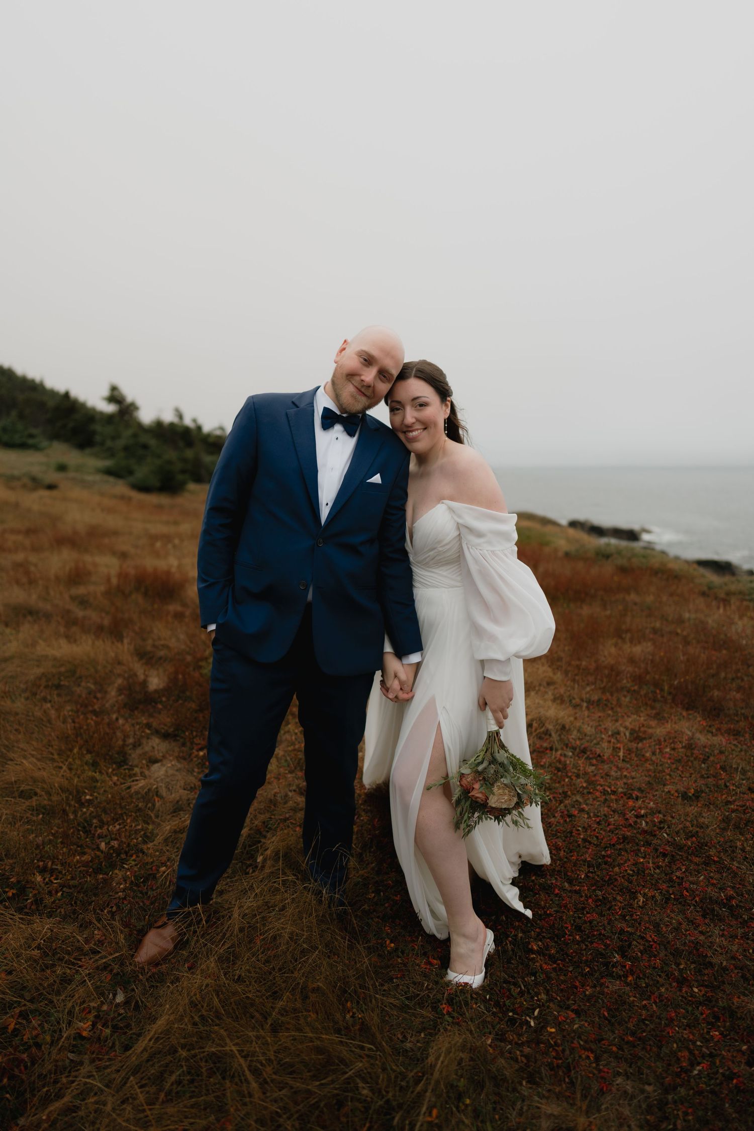 A couple in wedding attire shares intimate moments on a scenic coastal hillside at sunset.