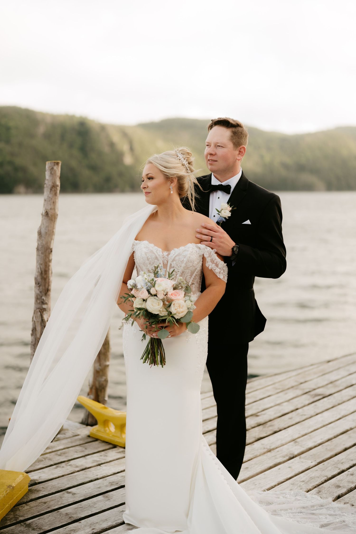 Bride and groom pose on a lakeside dock, bride in lace gown with bouquet, groom in black tuxedo, veil flowing in breeze.
