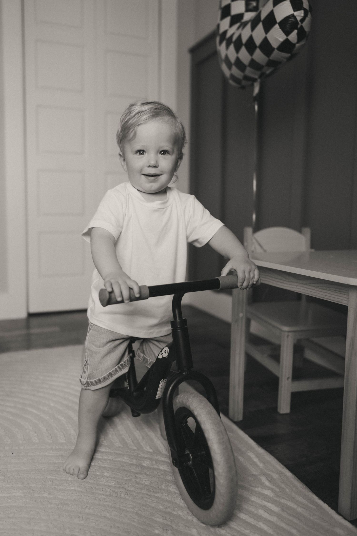 Toddler boy standing with a balance bike indoors in black and white photo with checkered balloons.