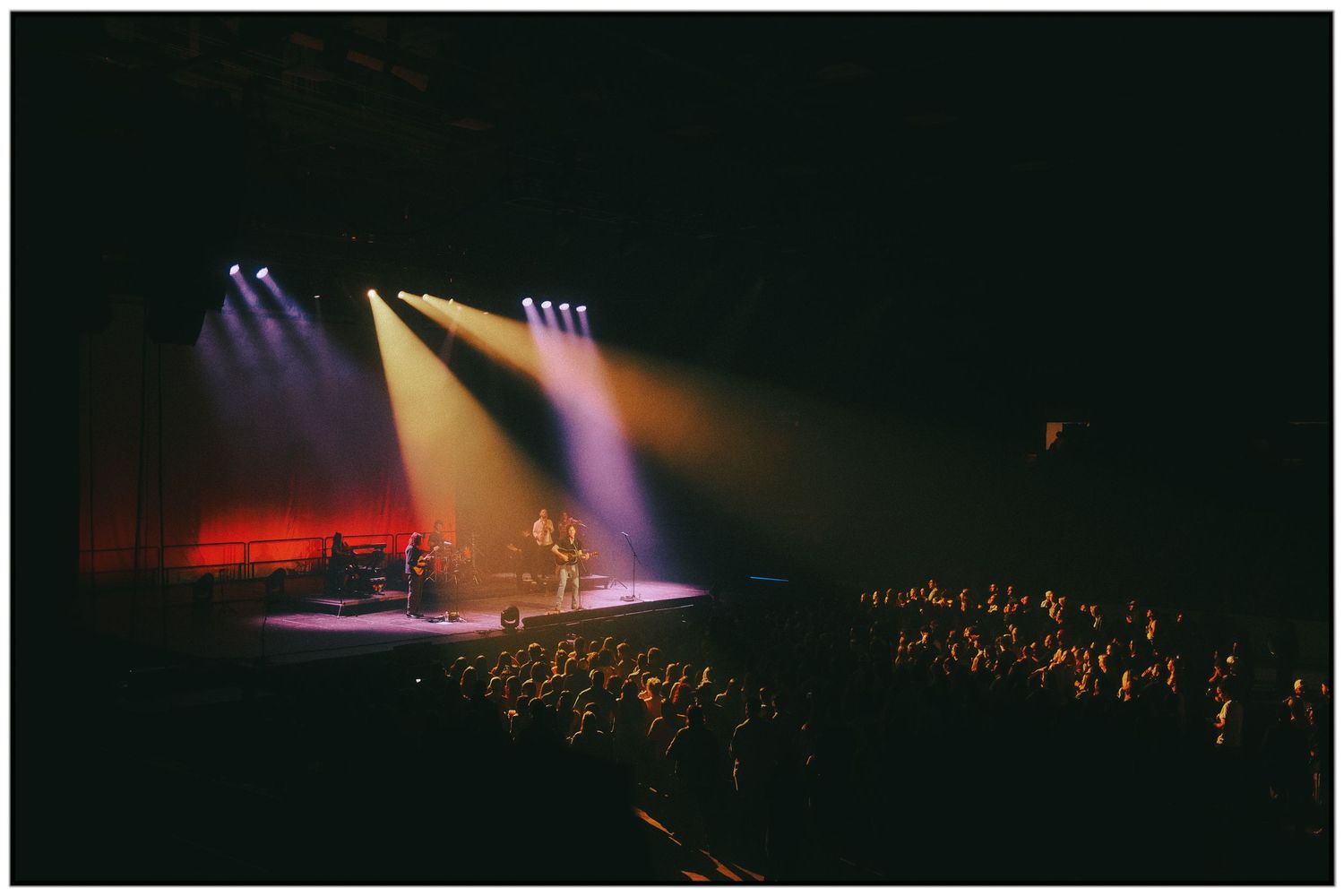Silhouetted performers on a dark stage illuminated by dramatic spotlights with a crowd visible in the foreground.