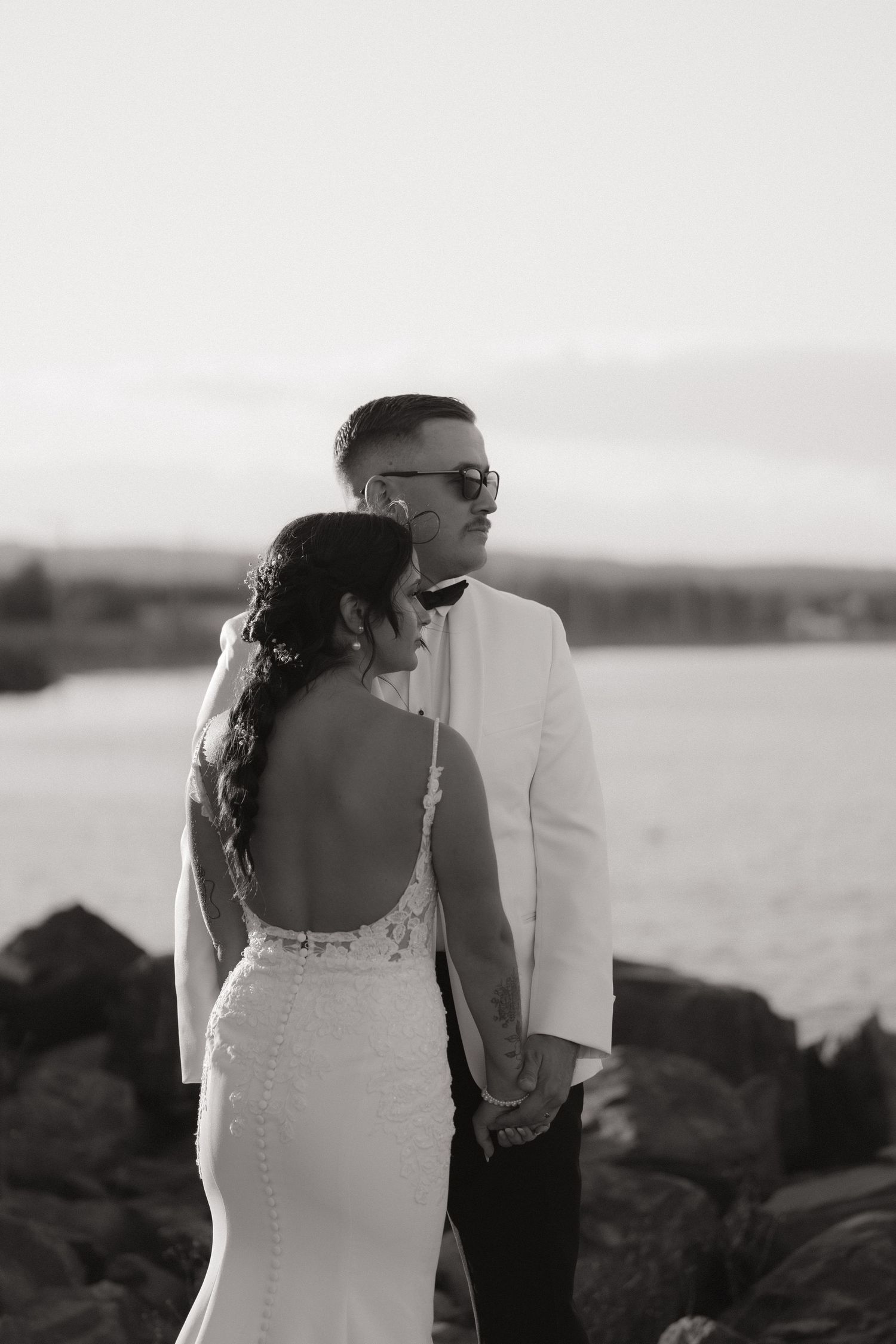 Black and white wedding photo of couple in formal attire standing by rocky shoreline at sunset.