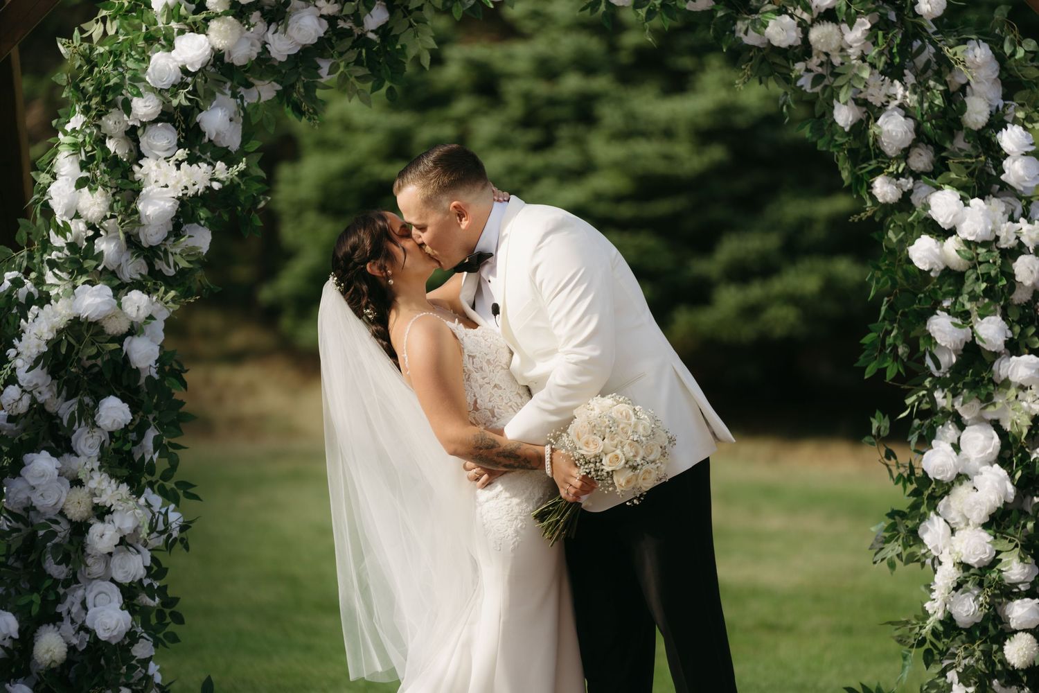 A romantic wedding couple shares tender kisses under a white floral arch in a lush garden setting.