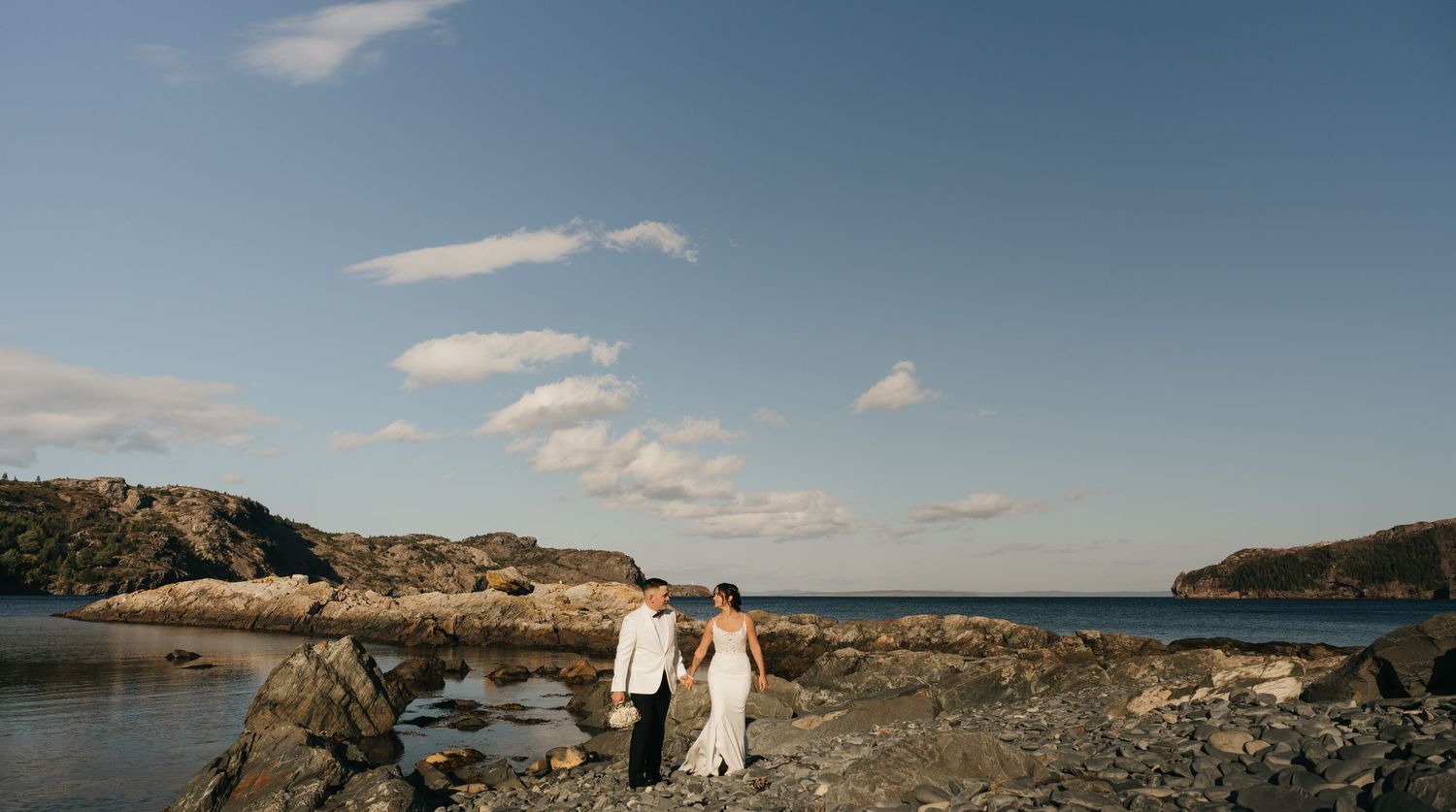 A panoramic coastal landscape shows rocky cliffs meeting the ocean under a bright blue sky with scattered clouds.