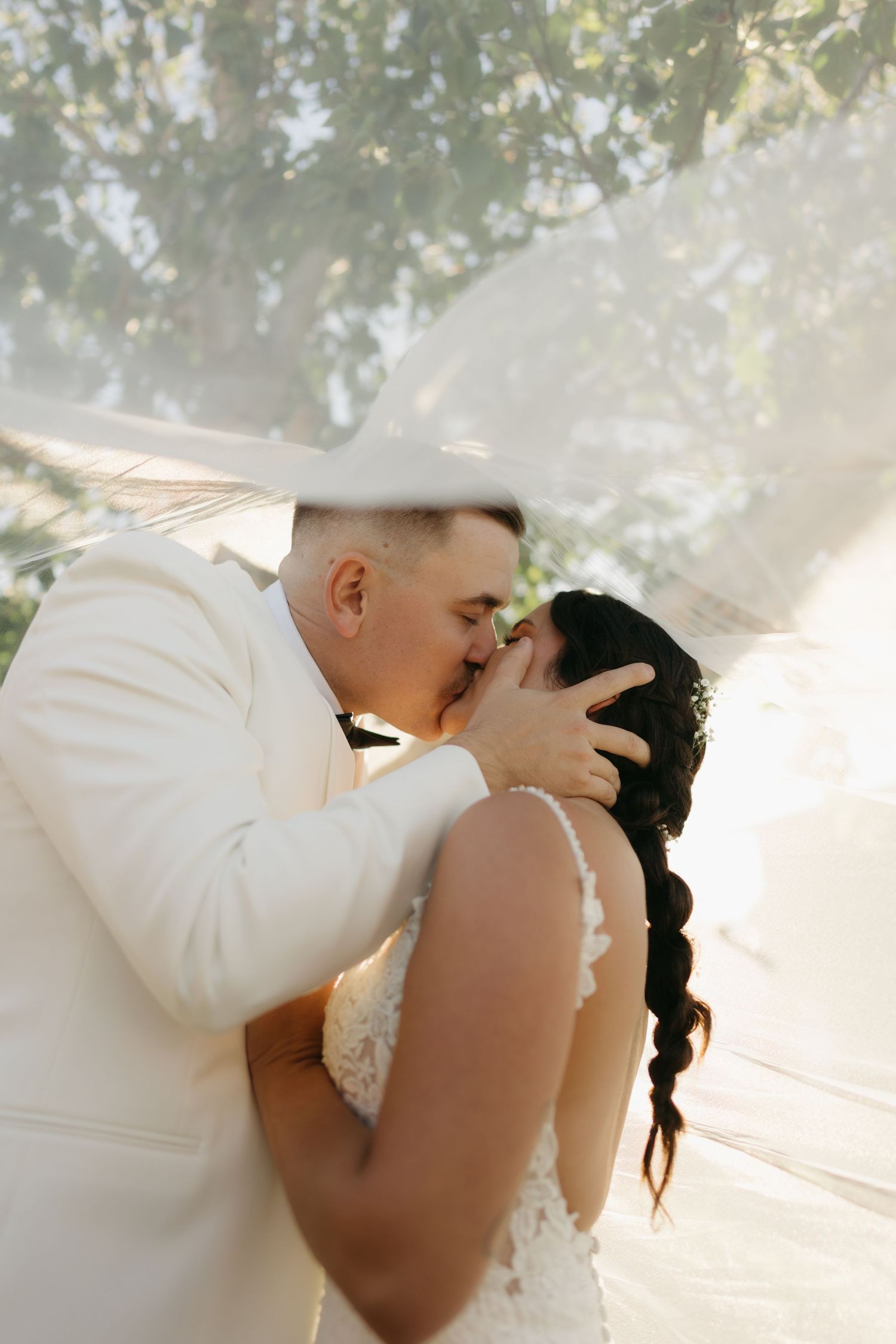 A romantic wedding couple shares a tender moment in soft sunlight beneath trees wearing white attire.