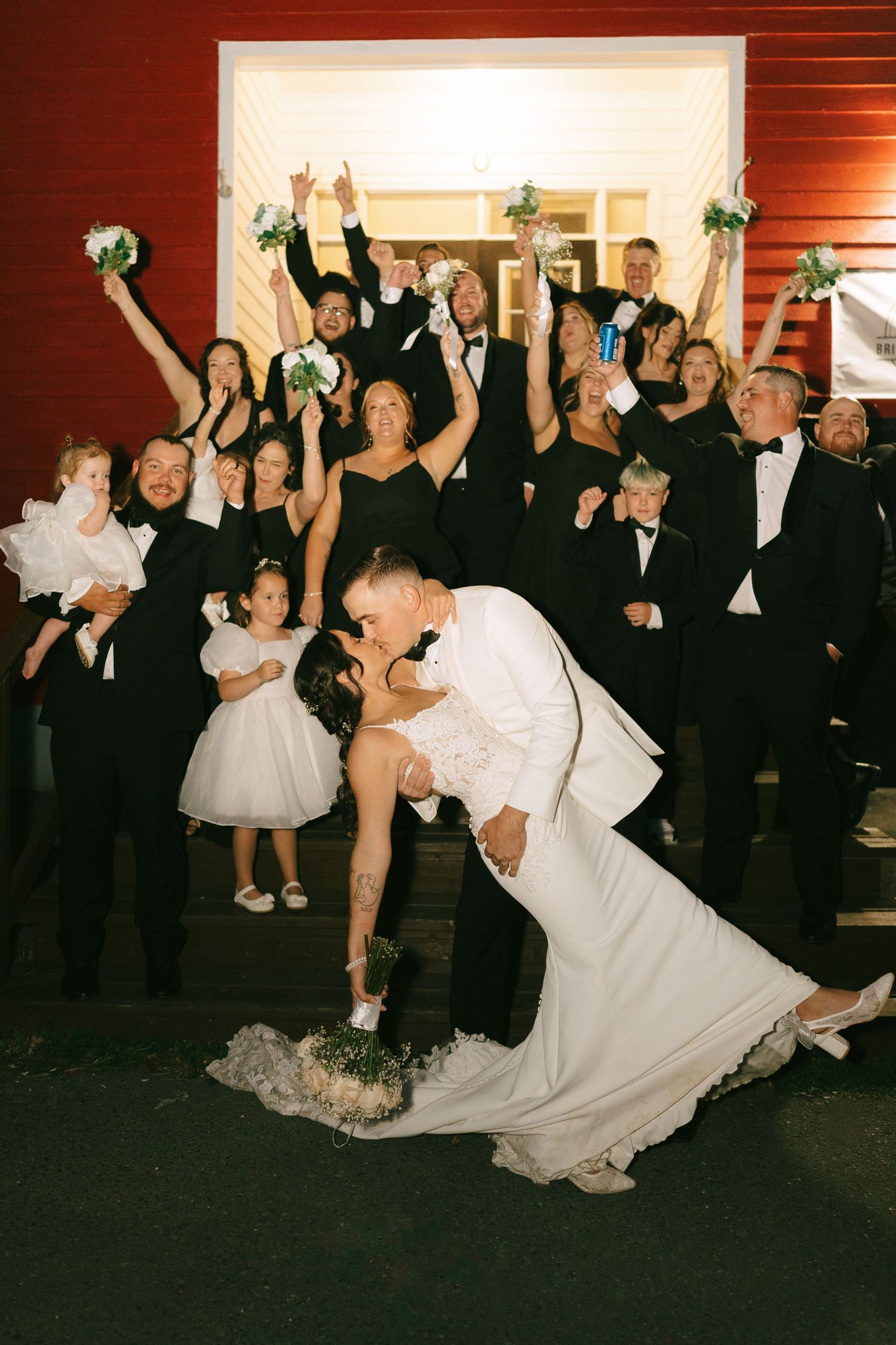 A wedding party celebrates as a couple shares a romantic dip kiss on the dance floor with guests cheering in the background.