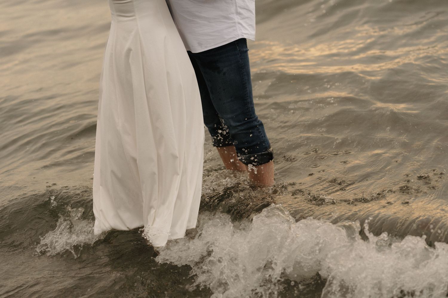 Newlywed couple walks along the beach shoreline as waves splash around their feet at sunset.