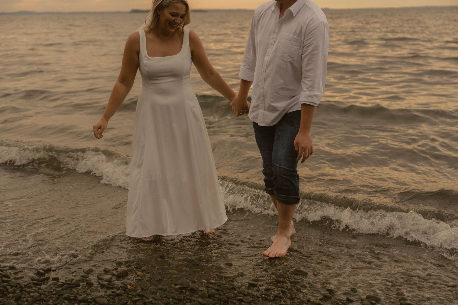 A couple in white and gray attire walks along the beach at sunset holding hands as waves roll in near the shore.