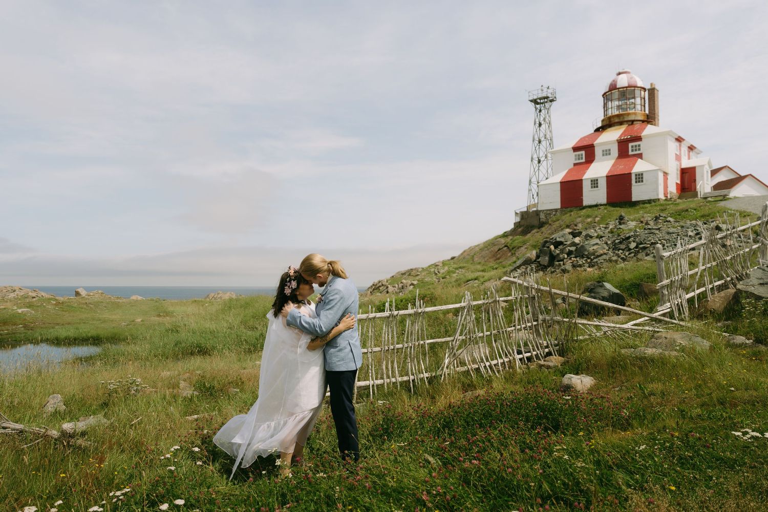 Couple sits on grassy hillside overlooking lighthouse and ocean at sunset in romantic coastal setting.