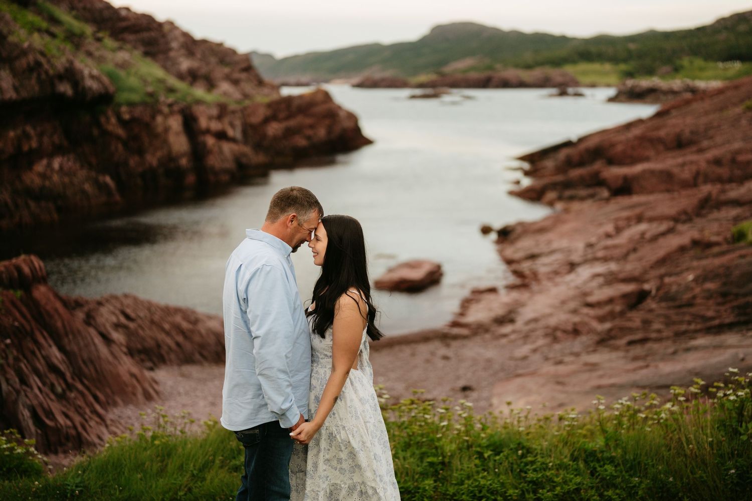 Couple embracing near red rock cliffs and a calm coastal inlet, surrounded by wildflowers and lush greenery.