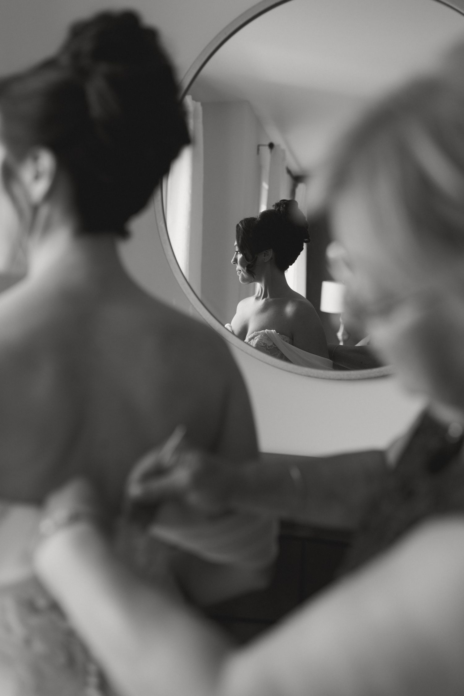Black and white photo of bride being helped into wedding dress by bridesmaids, reflected in round mirror.