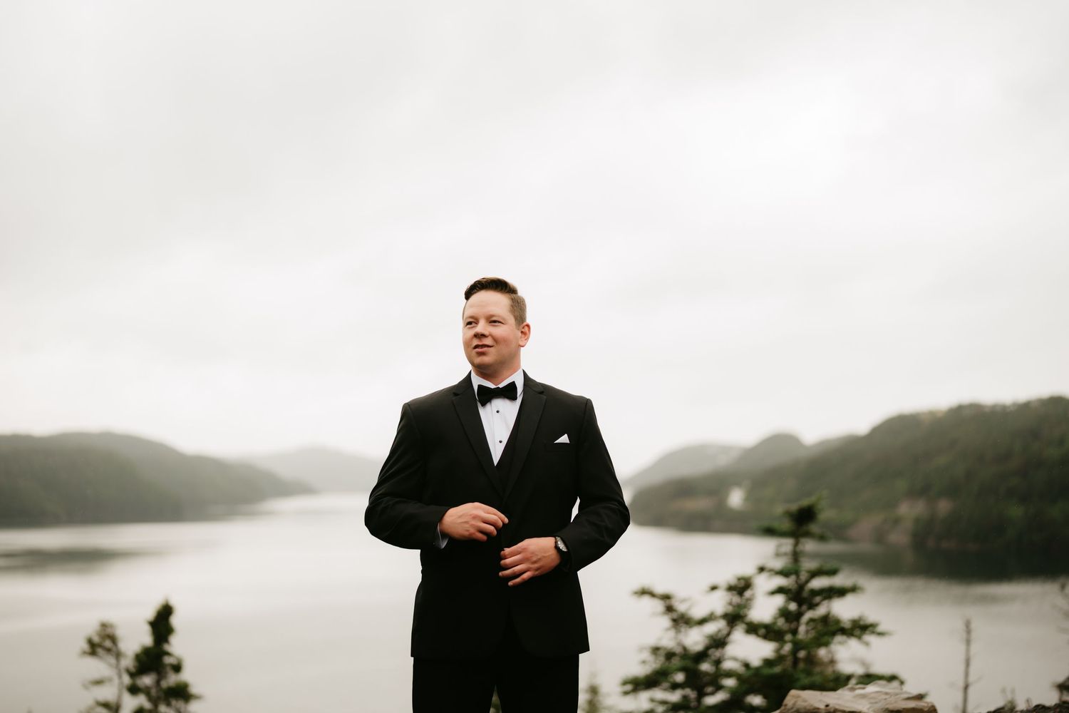 Groom in black tuxedo and bow tie stands before misty lake and mountain backdrop on overcast wedding day.