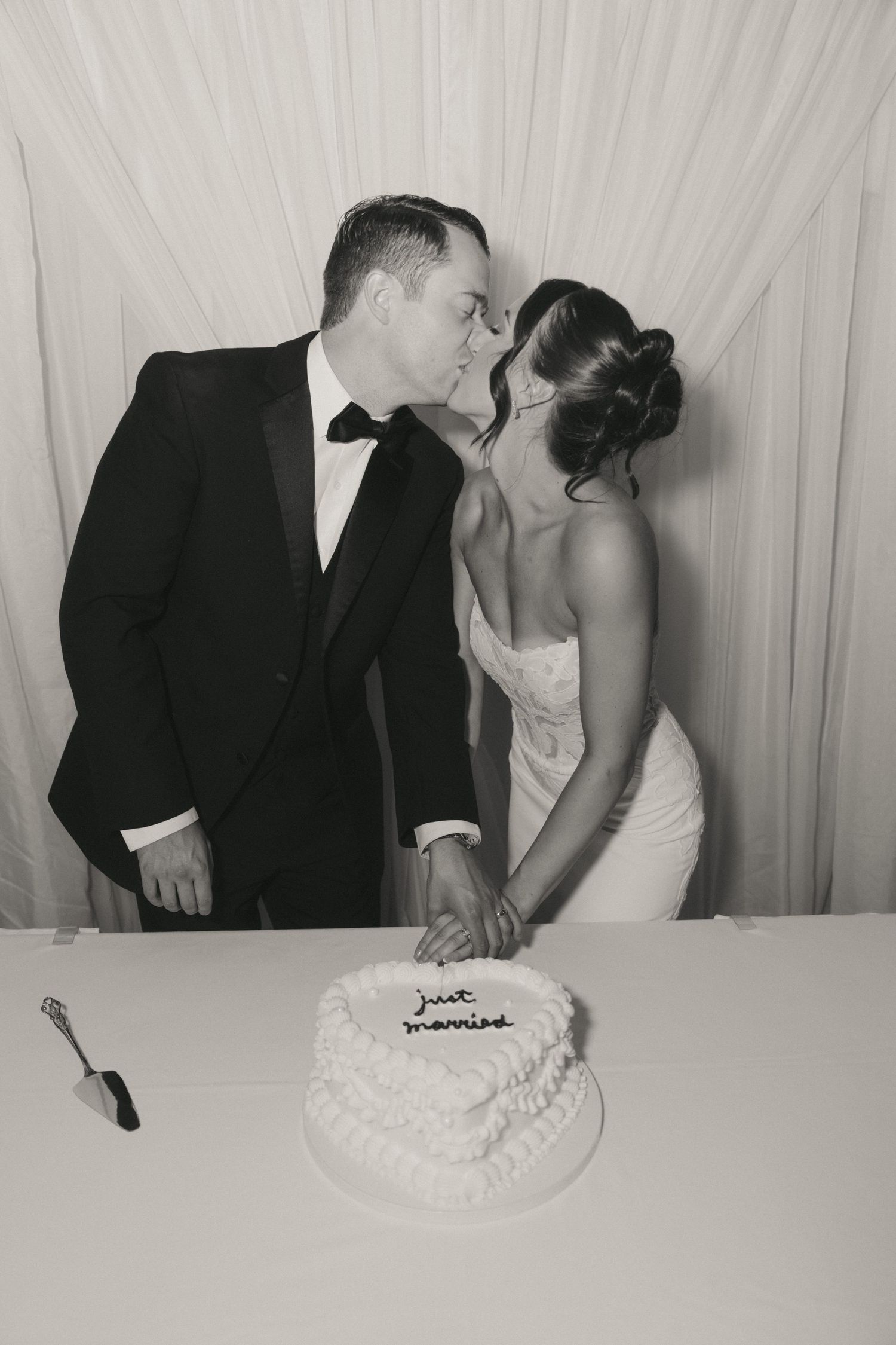 Black and white photo of newlyweds kissing while cutting their heart-shaped Just Married wedding cake.