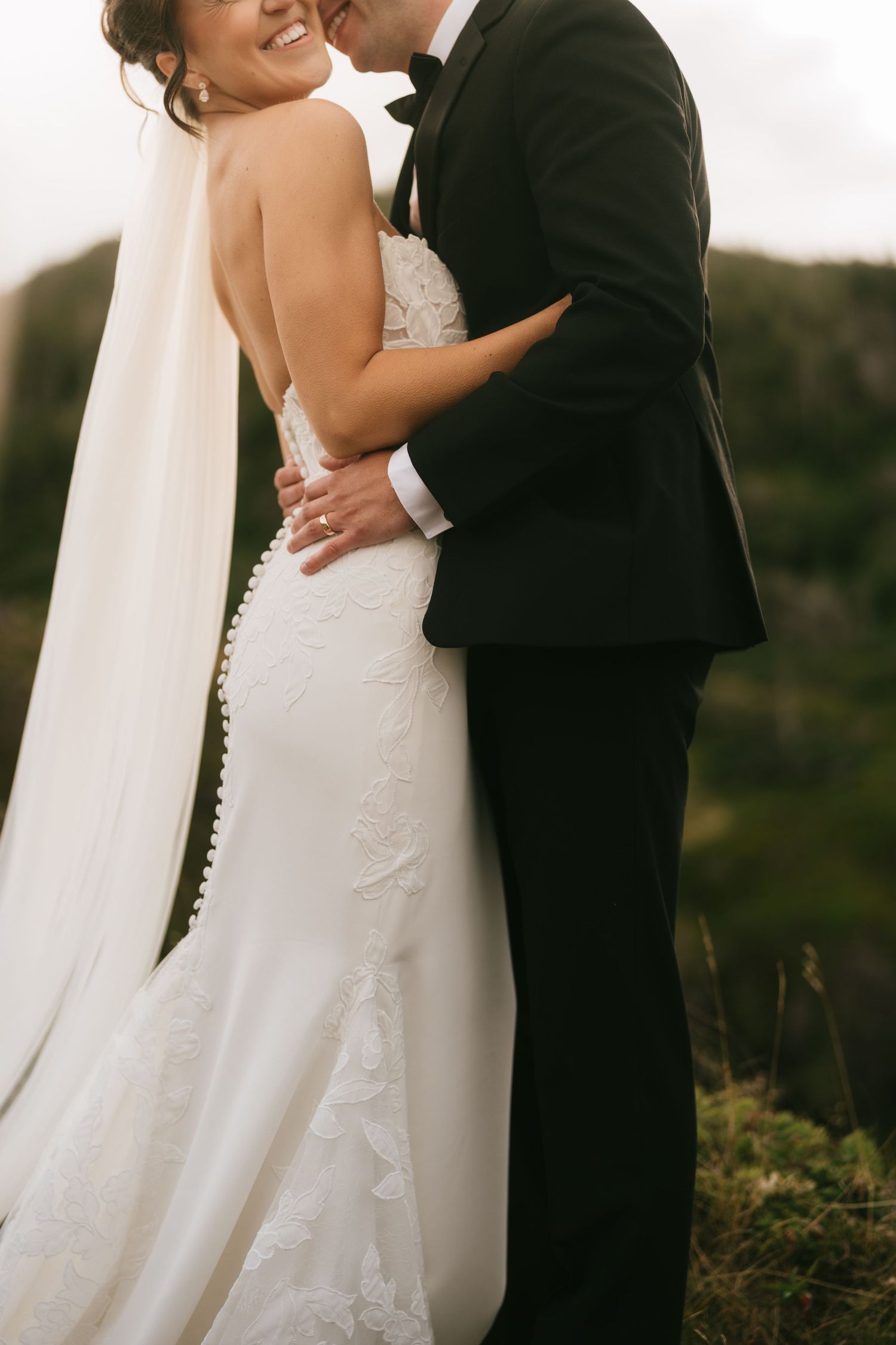 A bride in a lace mermaid gown and veil embraces her groom in a black tuxedo outdoors.