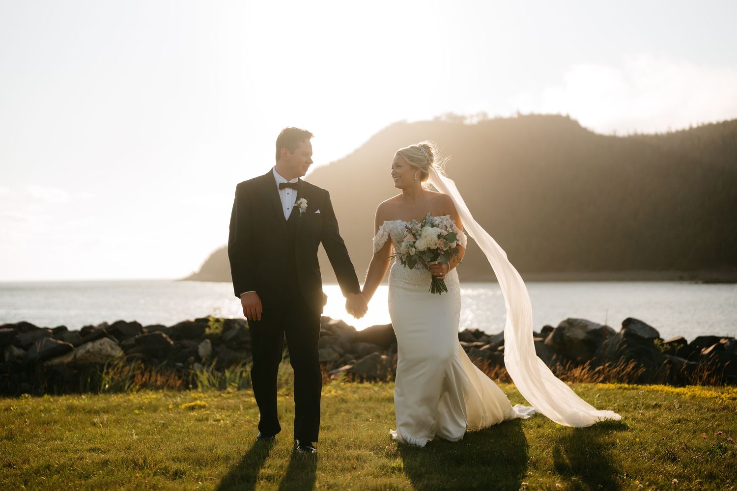 Bride and groom holding hands walking by the ocean at sunset, bride in white gown with flowing veil.