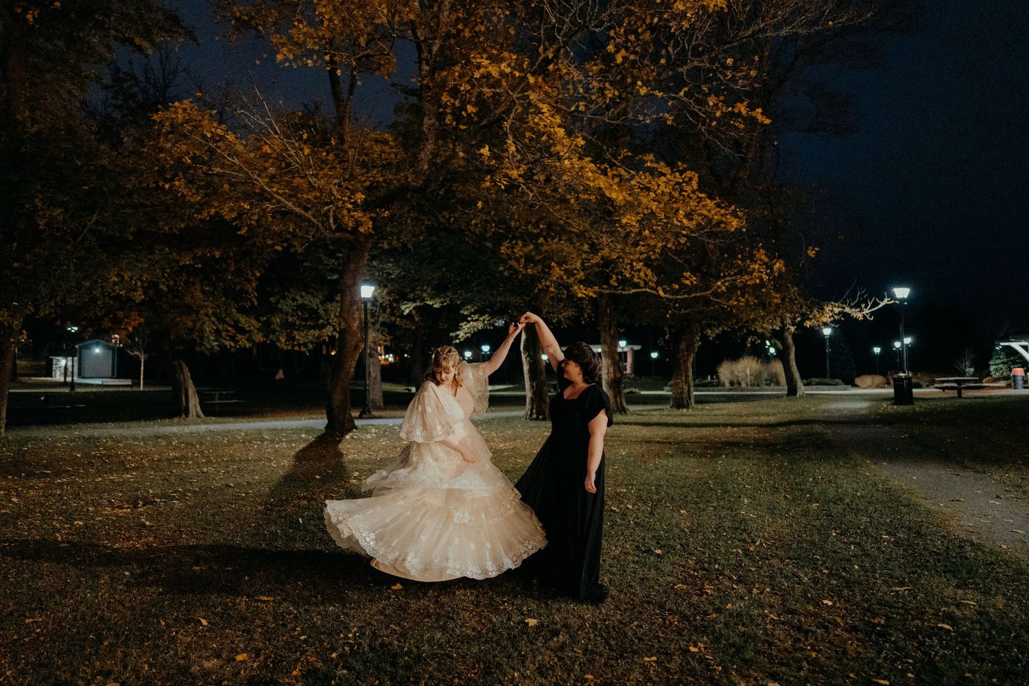 A couple in wedding attire walks through a park with autumn trees during golden hour lighting.