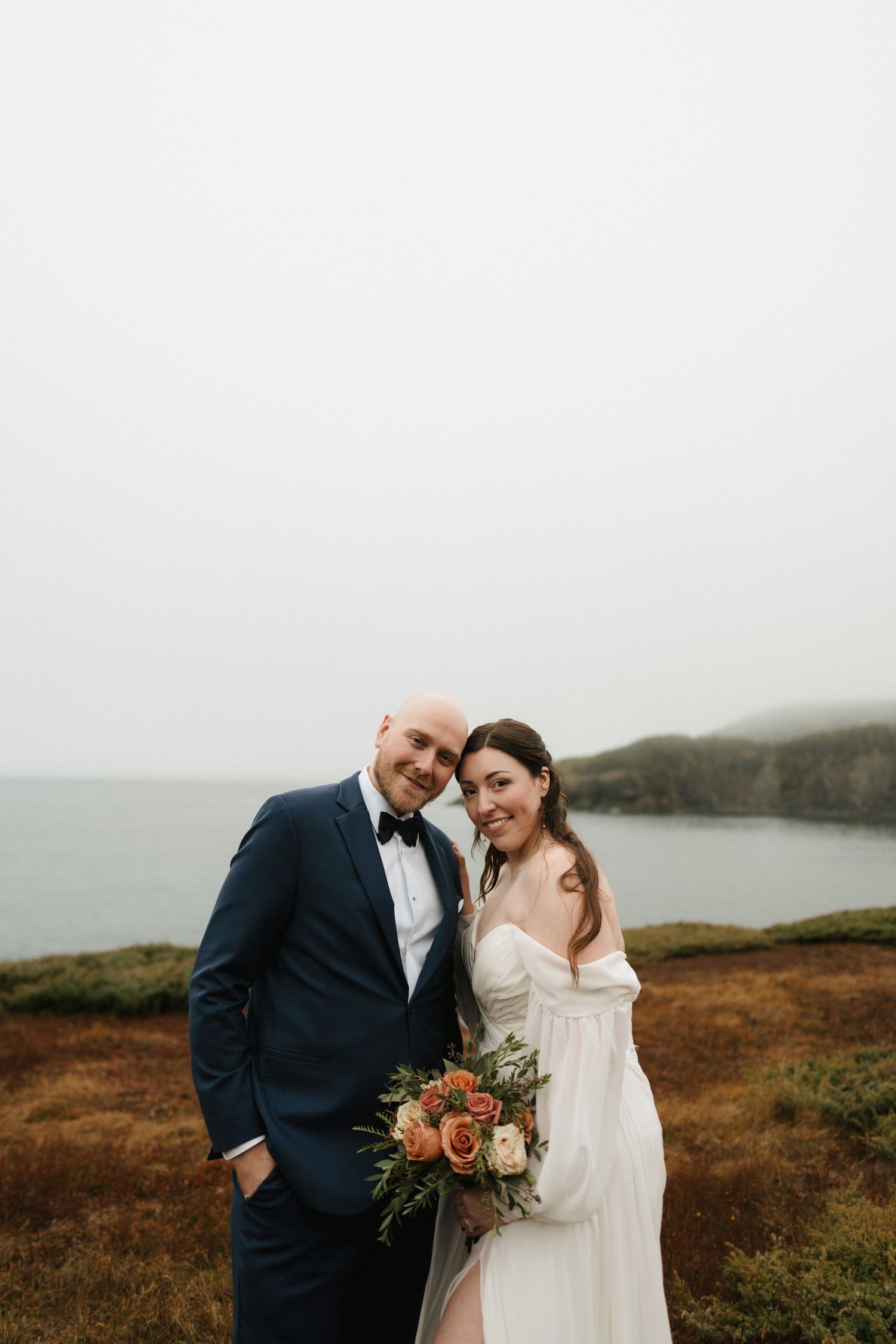 A couple in wedding attire share intimate moments against a misty coastal landscape with rolling hills.