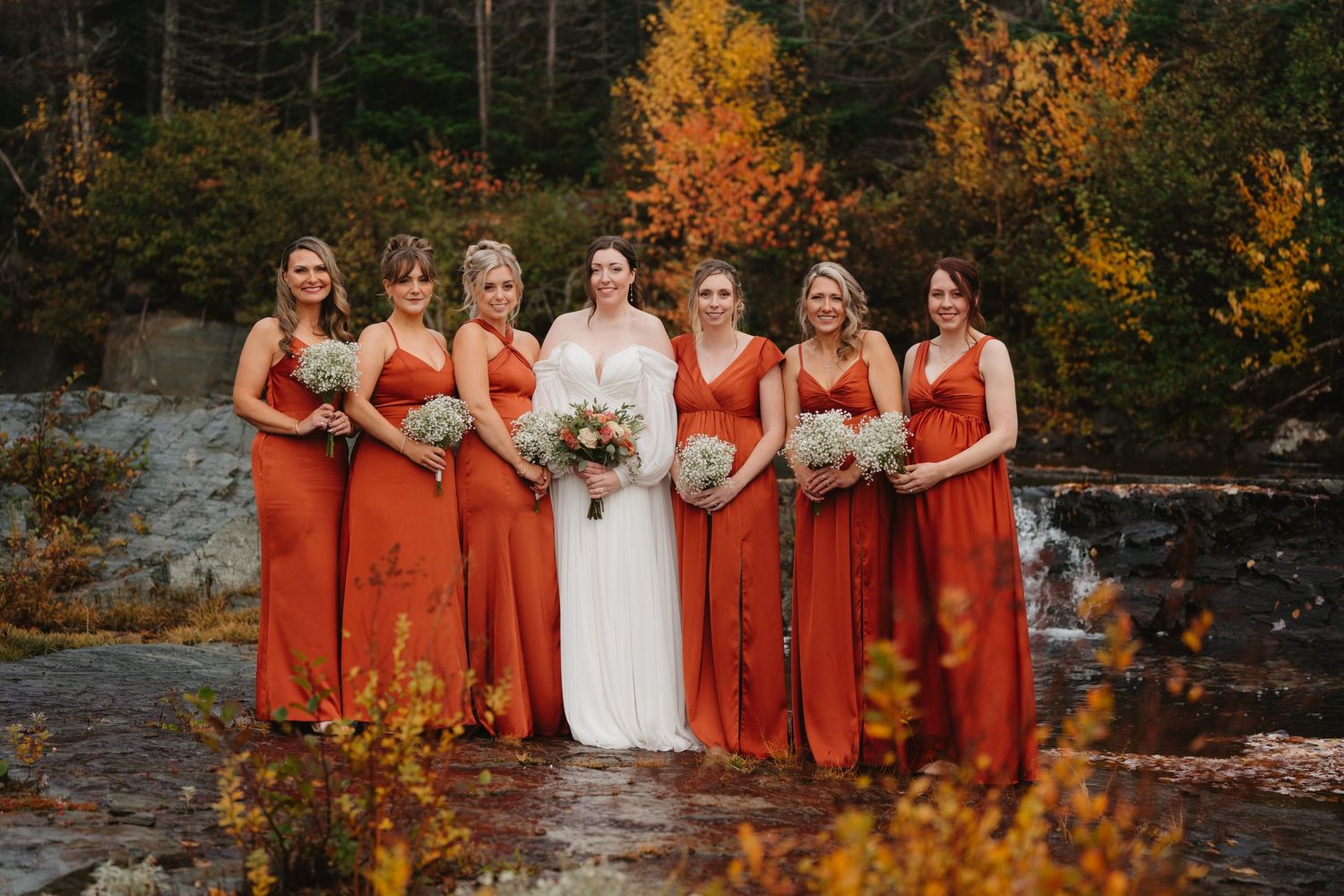A wedding party in burnt orange bridesmaid dresses poses against vibrant autumn foliage and stone walls.
