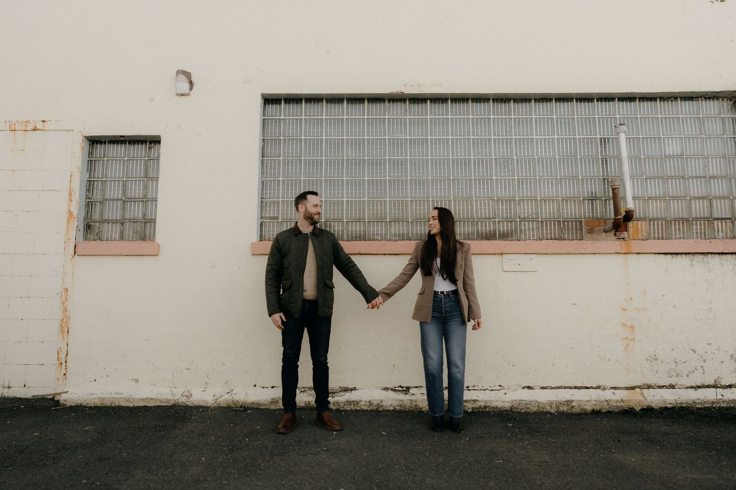 Couple holding hands facing each other against a white industrial building wall outdoors.