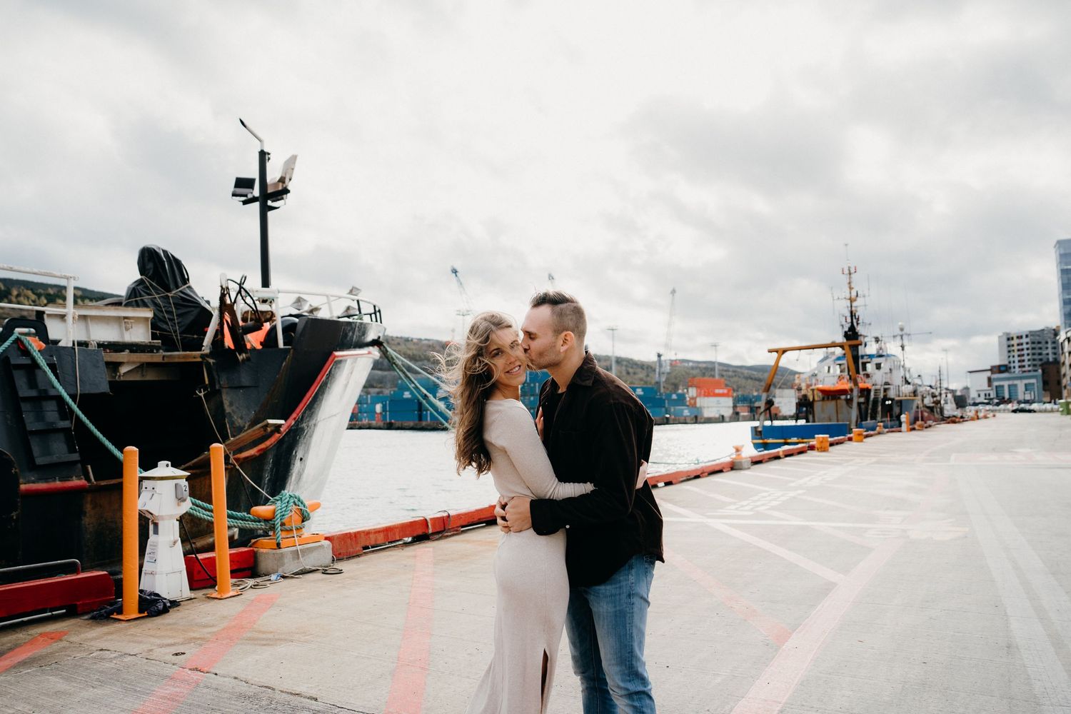 A couple stands close together on a harbor dock with moored vessels behind them.