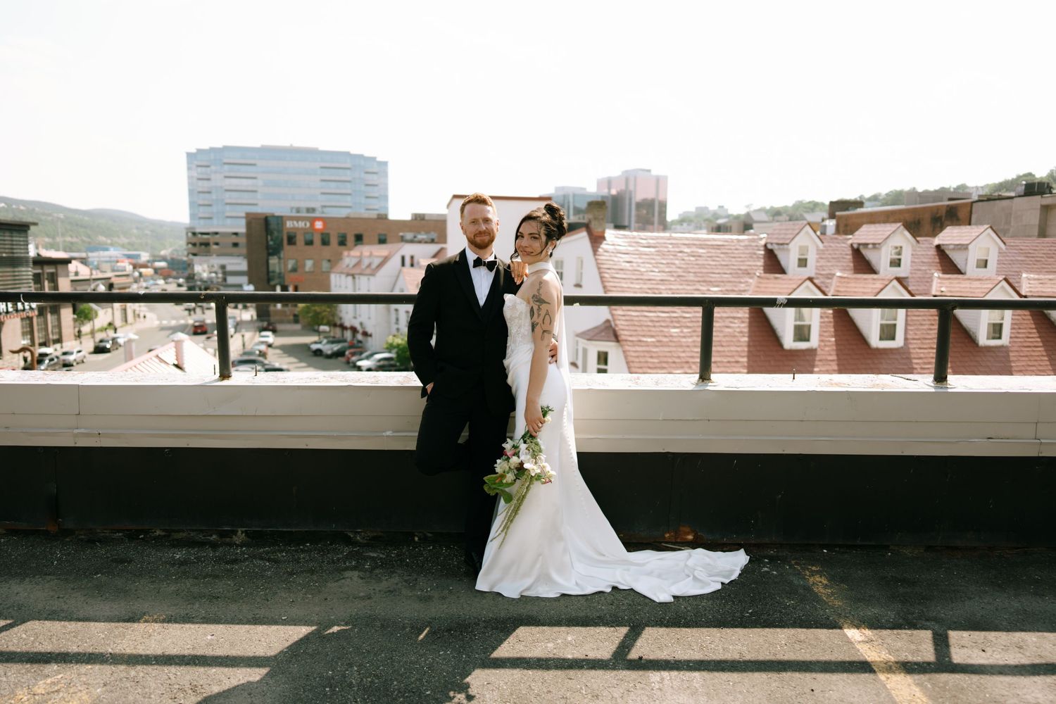 Bride and groom pose on rooftop terrace in elegant wedding attire with city skyline backdrop on sunny day.