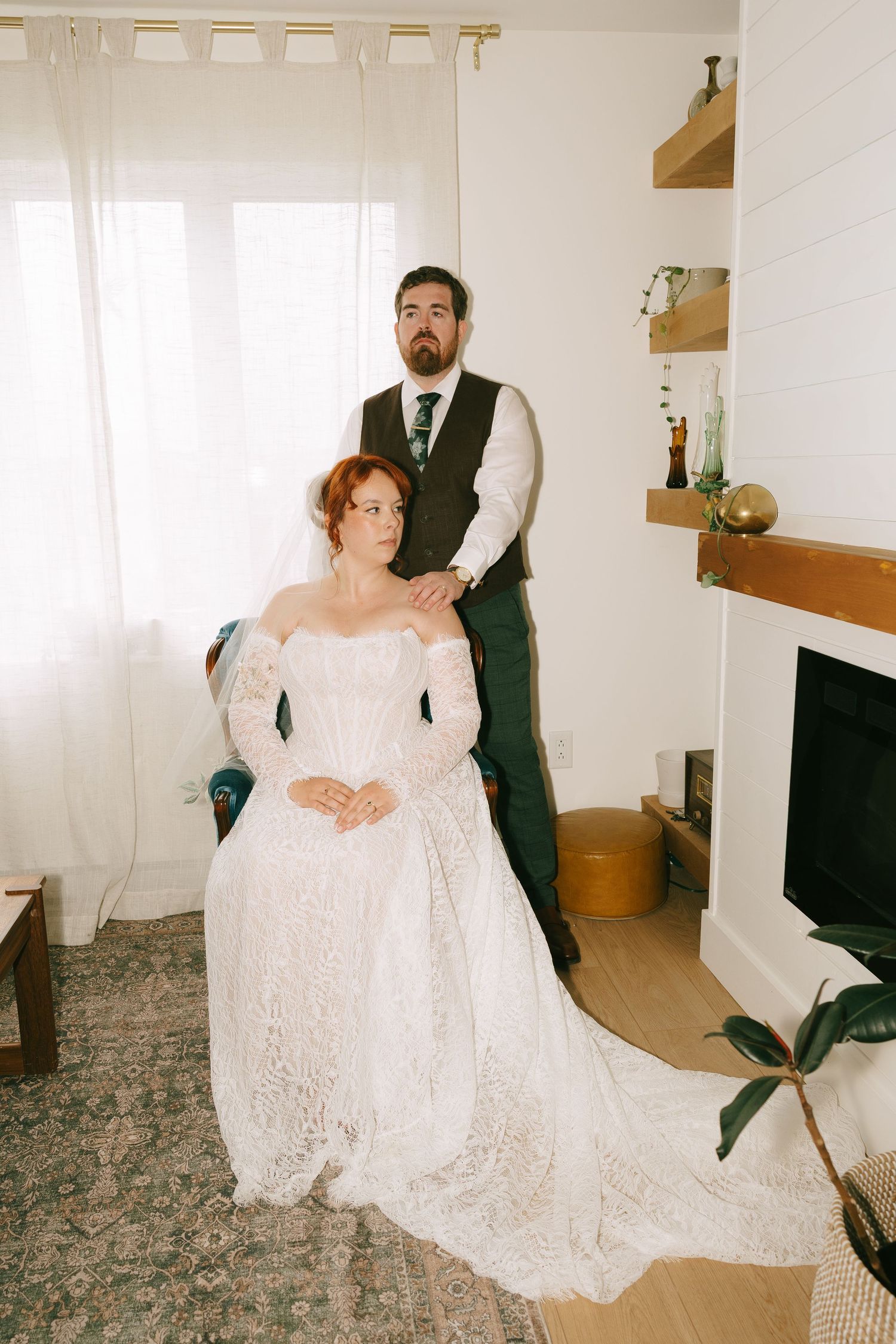 Bride in lace wedding gown seated while groom in vest adjusts her veil in a cozy living room.