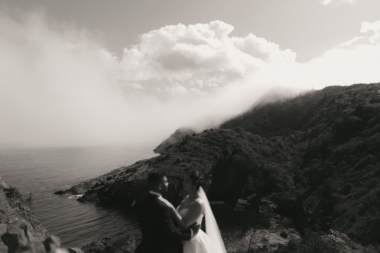Black and white photo of a couple embracing on a dramatic coastal cliff with misty mountains and ocean views.