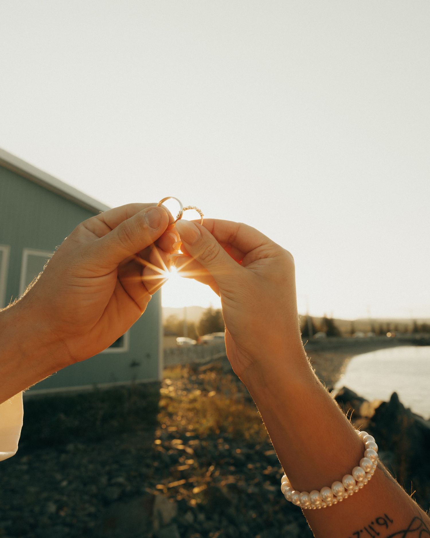 Hands holding a delicate pearl ring against a sunlit sky during golden hour.