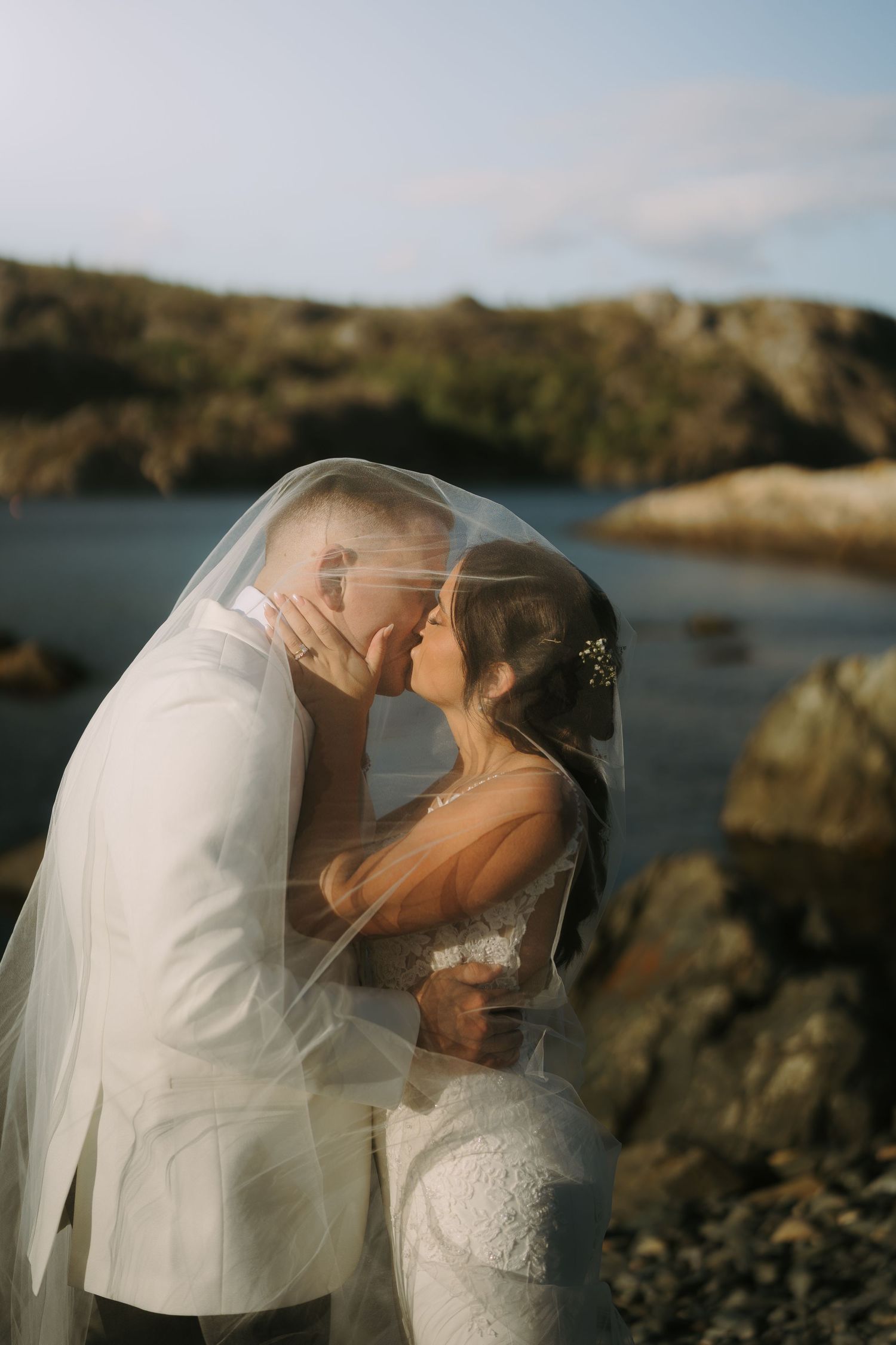 Romantic wedding couple shares a kiss under a flowing veil against a coastal backdrop with rocky cliffs at sunset.