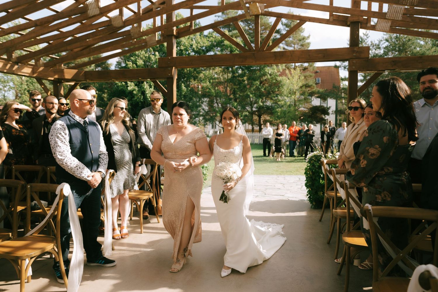 Guests seated in wooden chairs at an outdoor wedding ceremony under a rustic wooden pavilion structure.