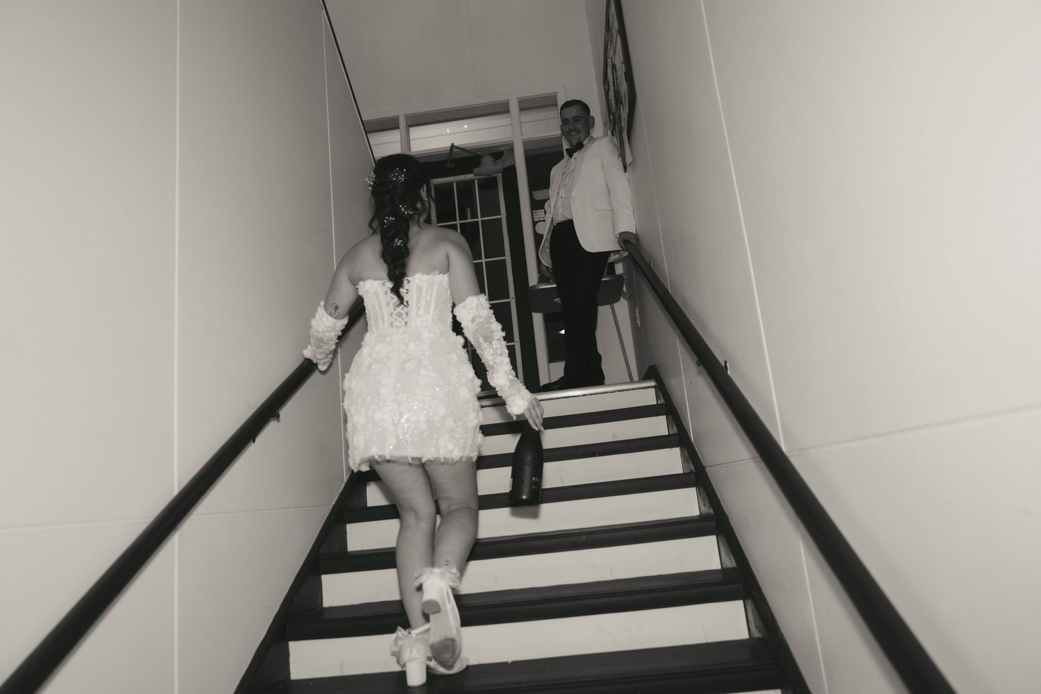 A black and white photograph shows someone walking up a stark staircase with dark handrails and white walls.