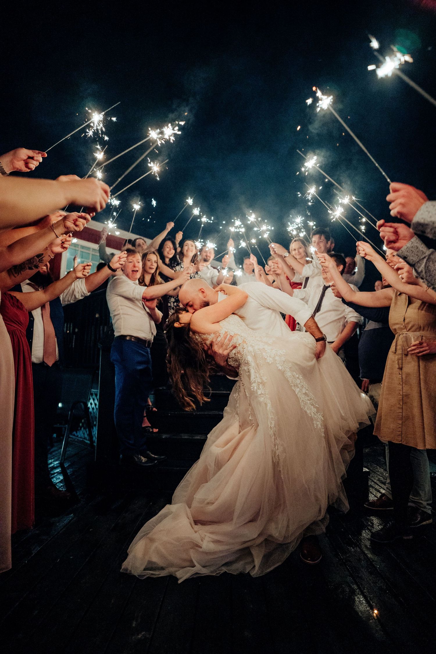 Wedding guests hold sparklers in a curved line formation creating a magical nighttime tunnel effect at an outdoor celebration.