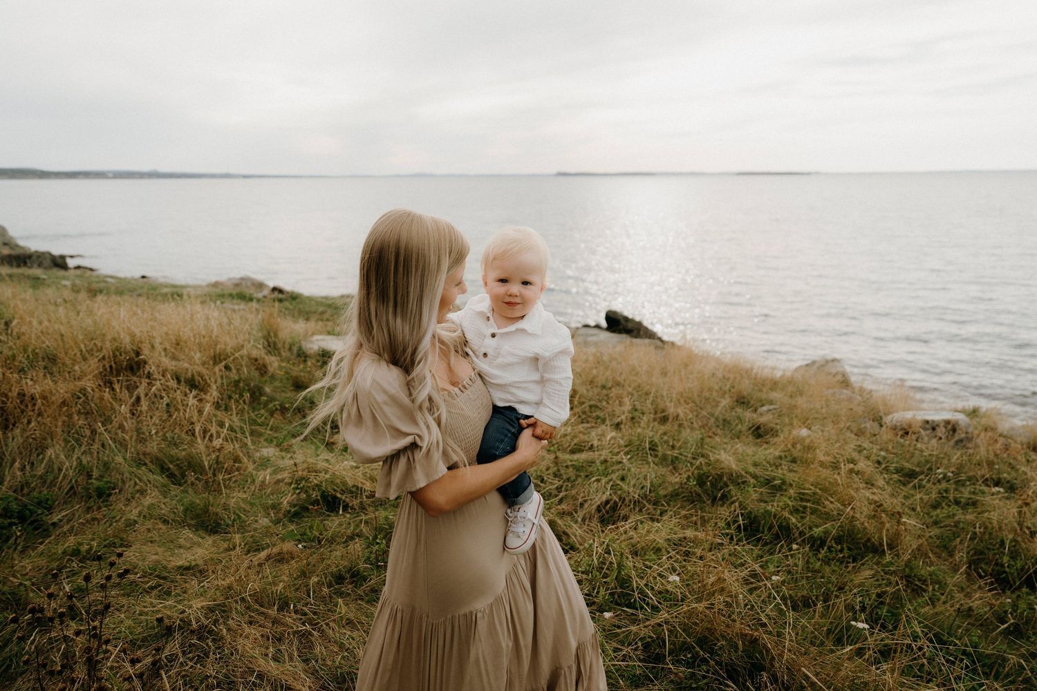 A parent and child stand together on a grassy coastal cliff overlooking the ocean on a cloudy day.