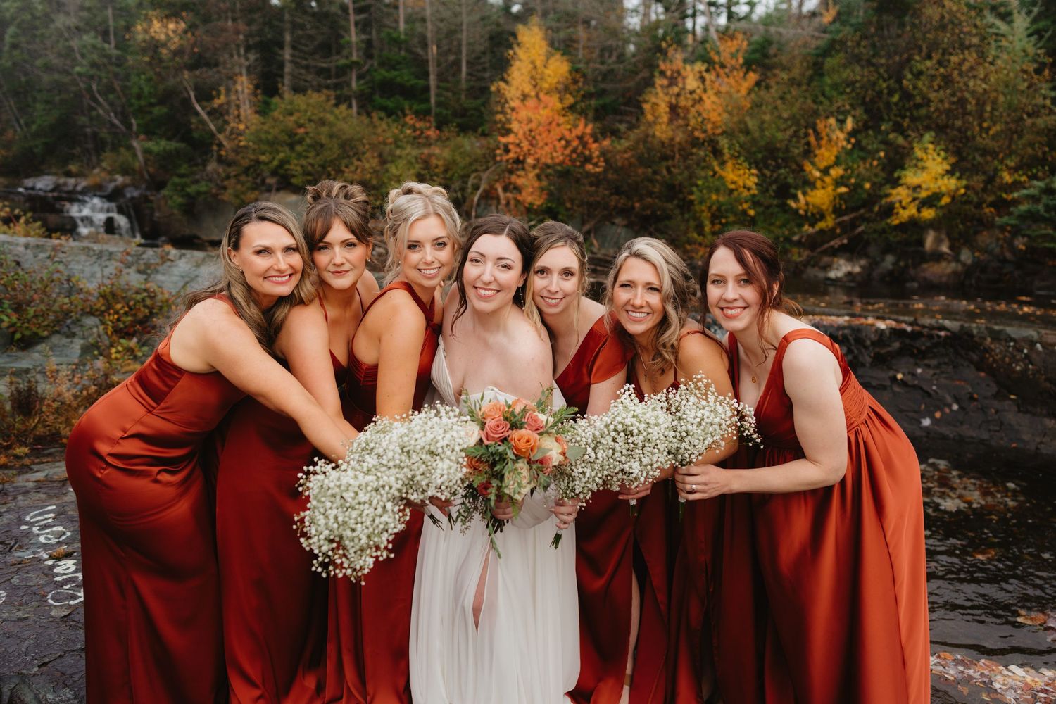 A bridal party in burgundy dresses poses together in a scenic autumn forest setting with colorful fall foliage.