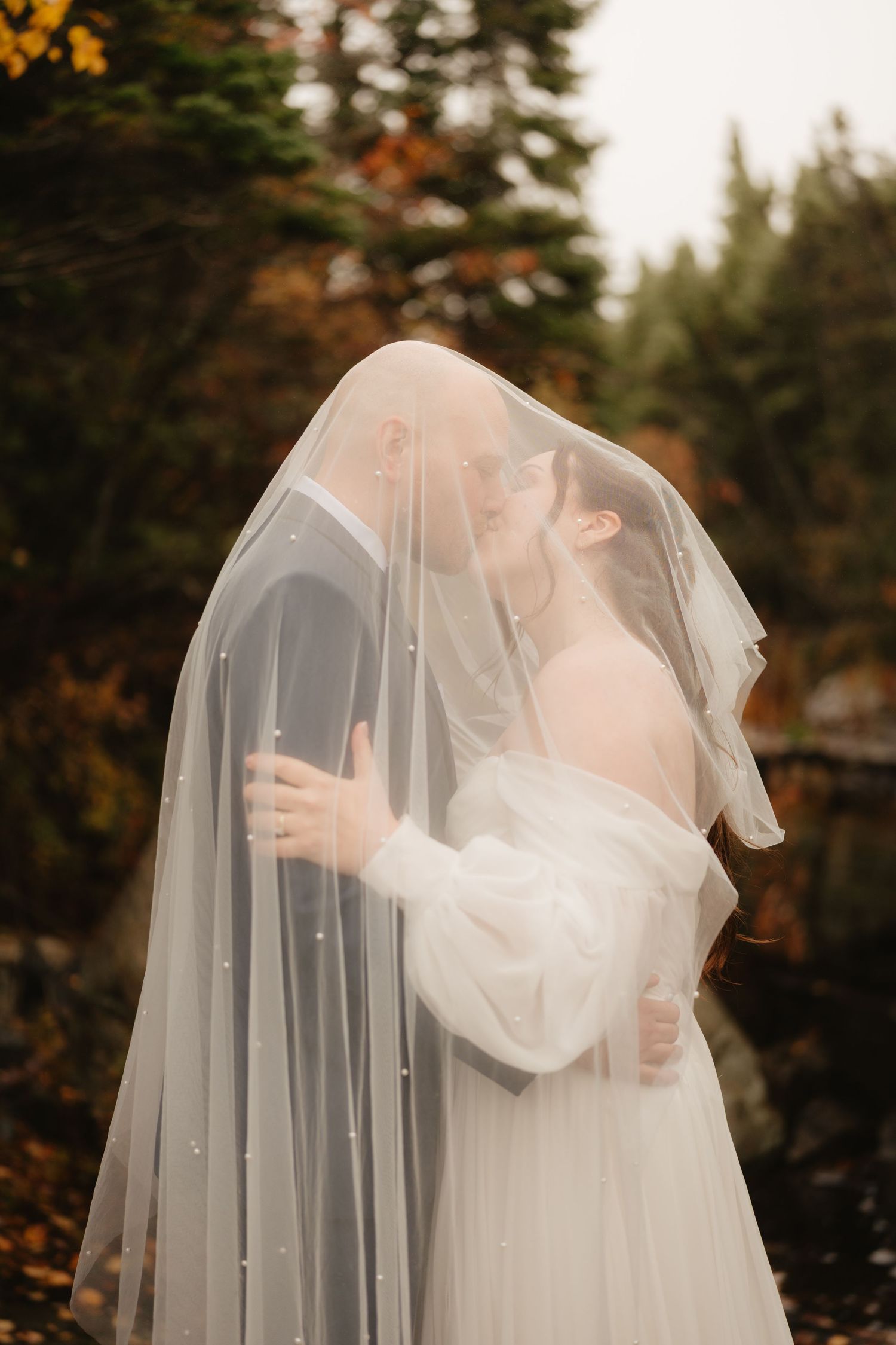 A romantic autumn wedding scene with a couple sharing an intimate moment under a delicate veil in the woods.
