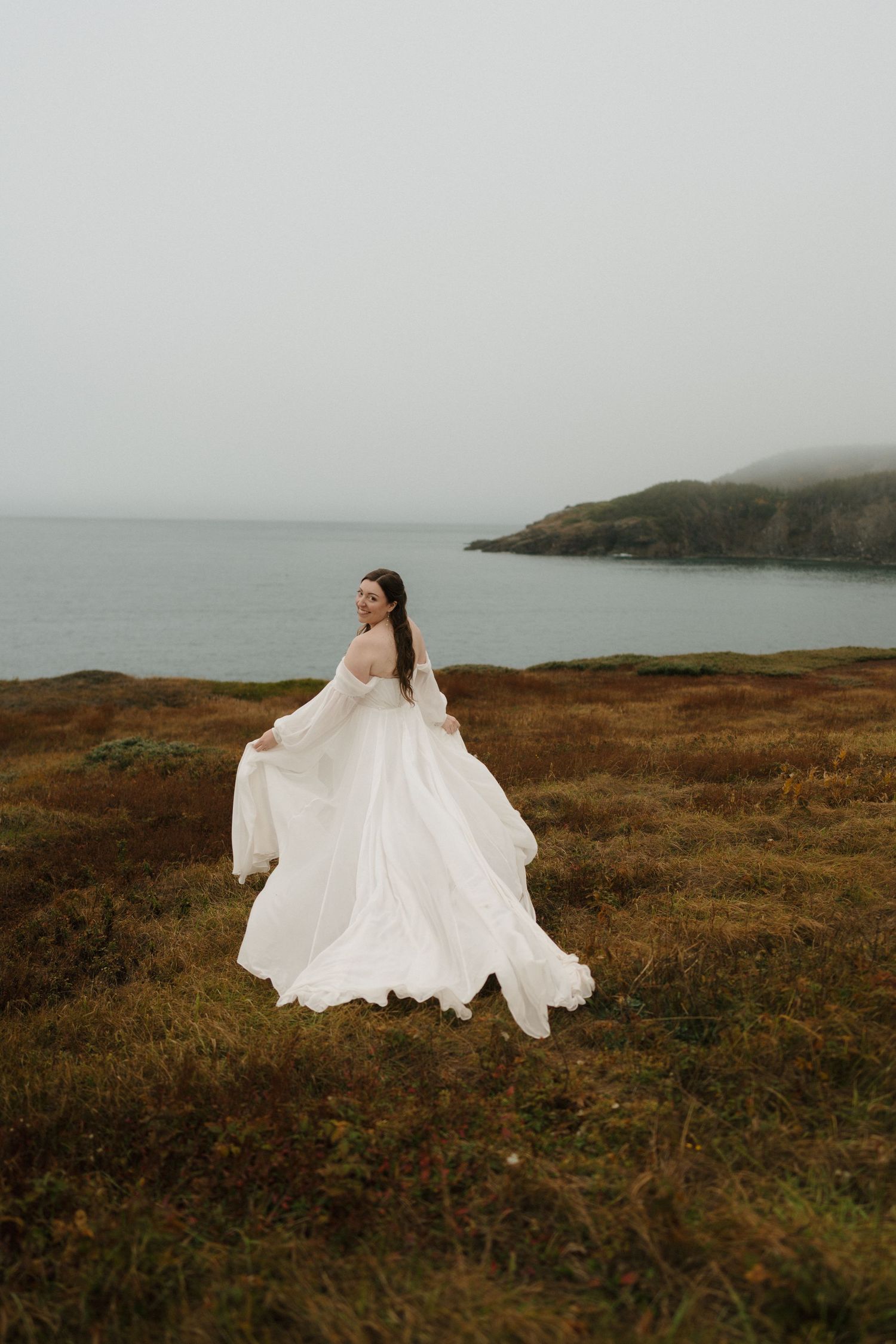 A flowing white wedding dress billows in the wind on a misty coastal cliff overlooking the moody sea.