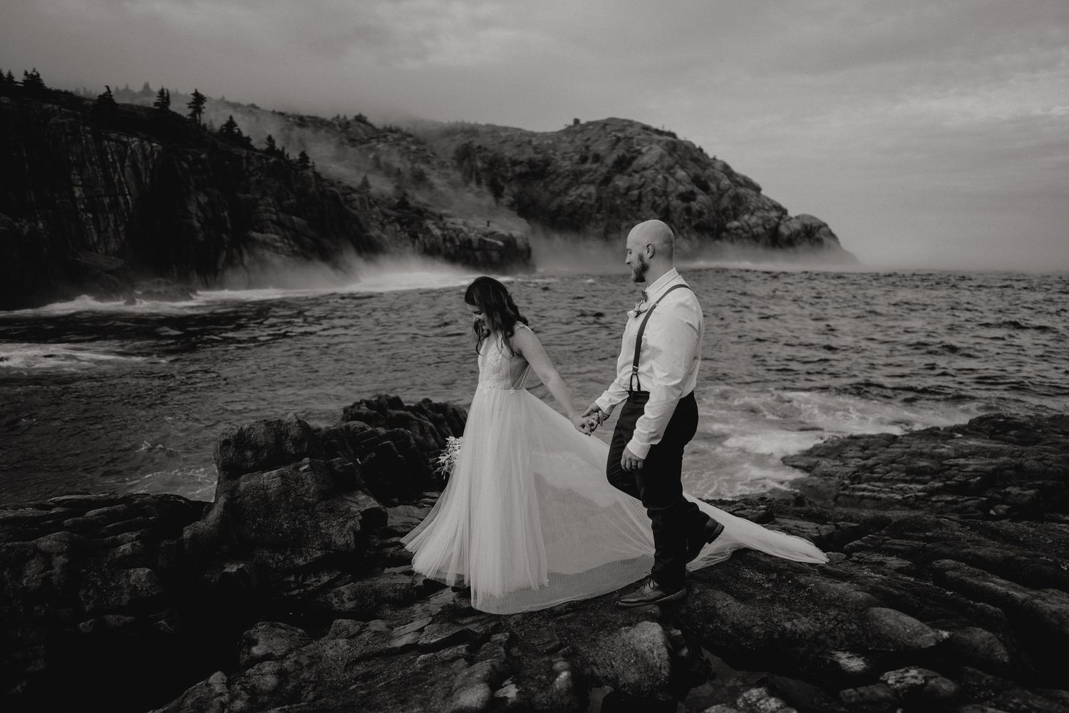 Couple walks along dramatic rocky coastline during black and white wedding photoshoot.