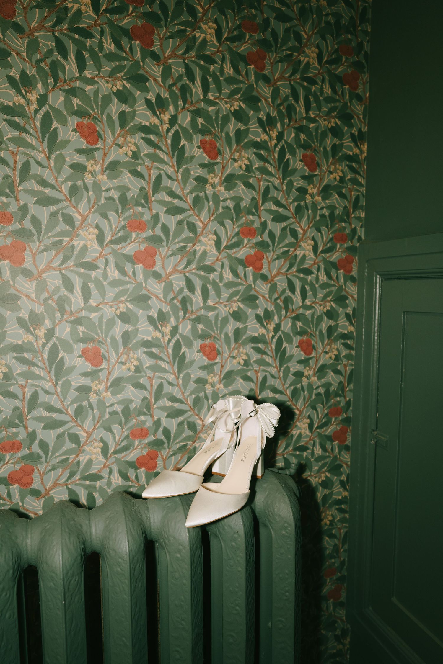 Elegant white heels arranged on top of a vintage green radiator with floral wallpaper.