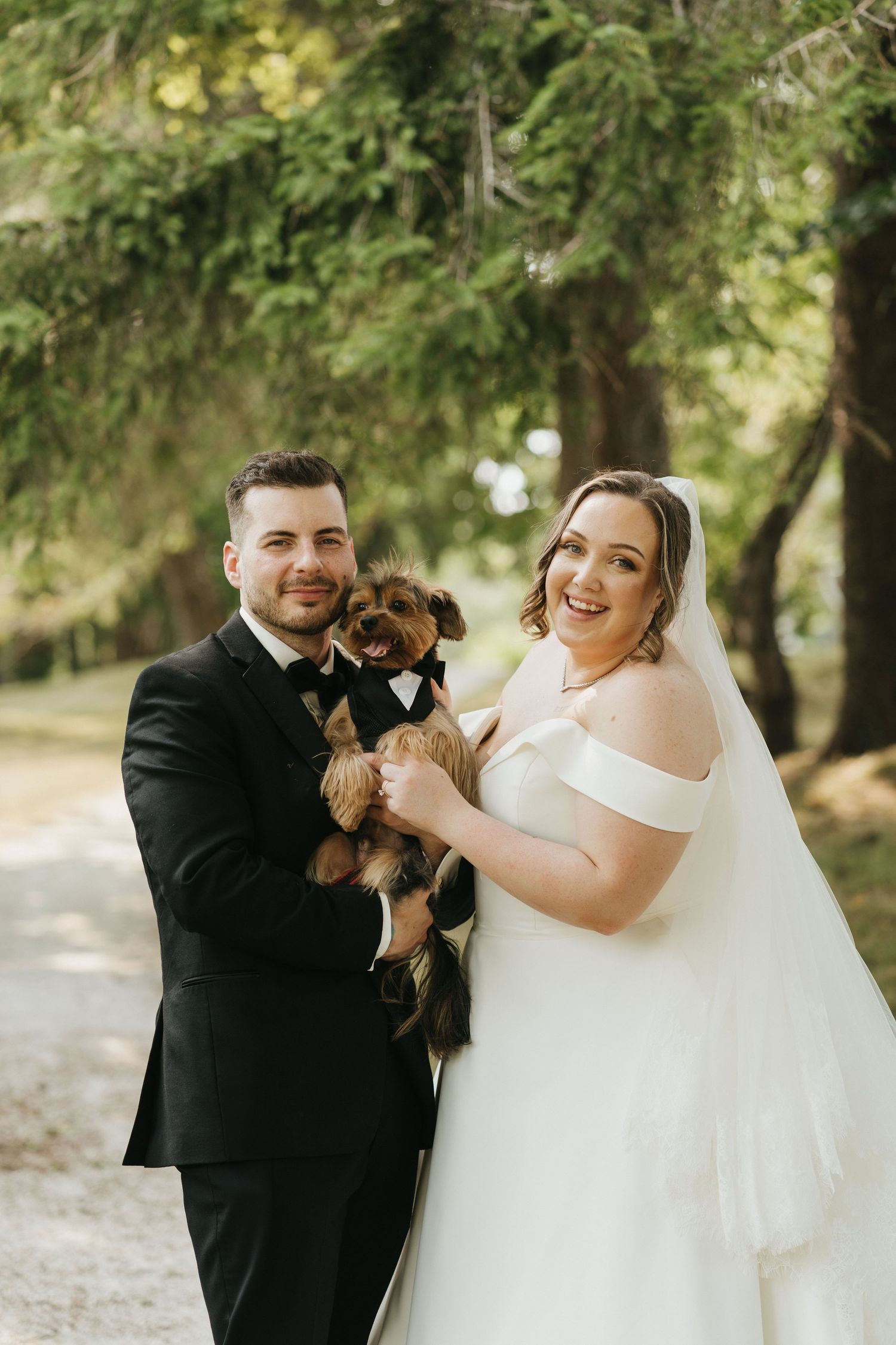 Happy couple in wedding attire holds their small dog while posing in a park.