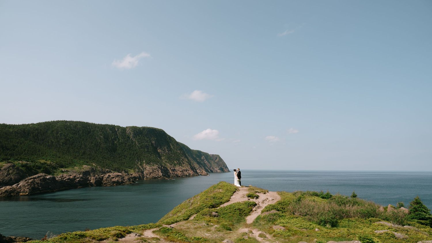 A couple stands on a scenic coastal cliff overlooking turquoise waters and lush green hills under a clear blue sky.