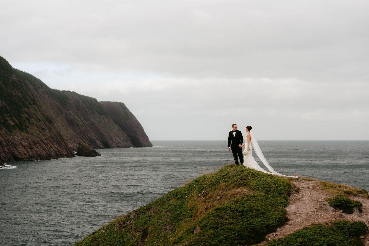 Bride and groom stand on a dramatic coastal cliff overlooking the ocean on a cloudy day, with the bride's veil flowing in the wind.