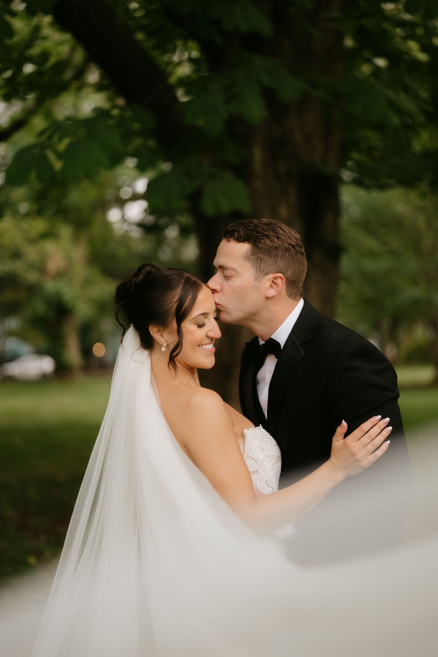 Groom in black tuxedo kissing bride on forehead outdoors, bride wearing lace gown and flowing veil near green trees.