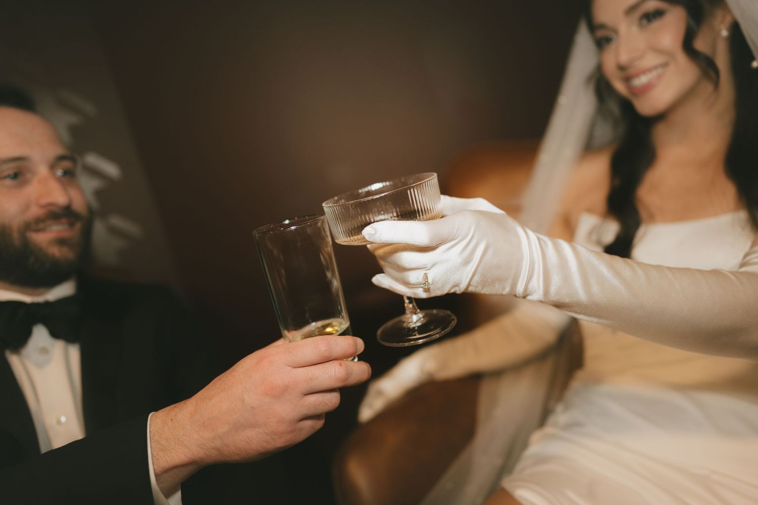 Bride in white gloves and groom in tuxedo clinking champagne glasses during wedding toast celebration.