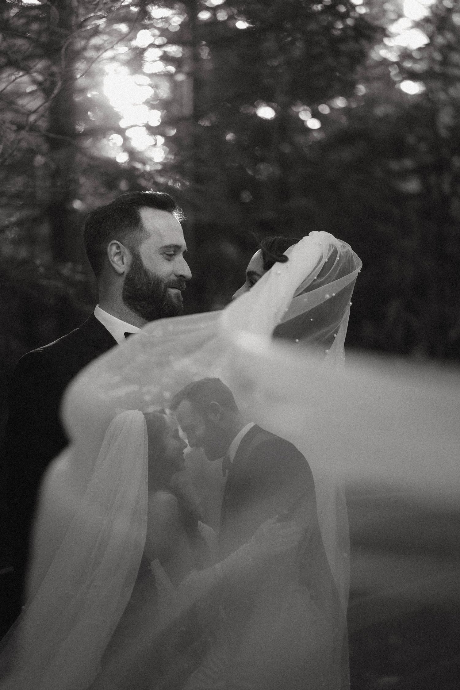 Black and white wedding photo of bride and groom embracing, veil flowing, surrounded by trees with soft bokeh lighting.