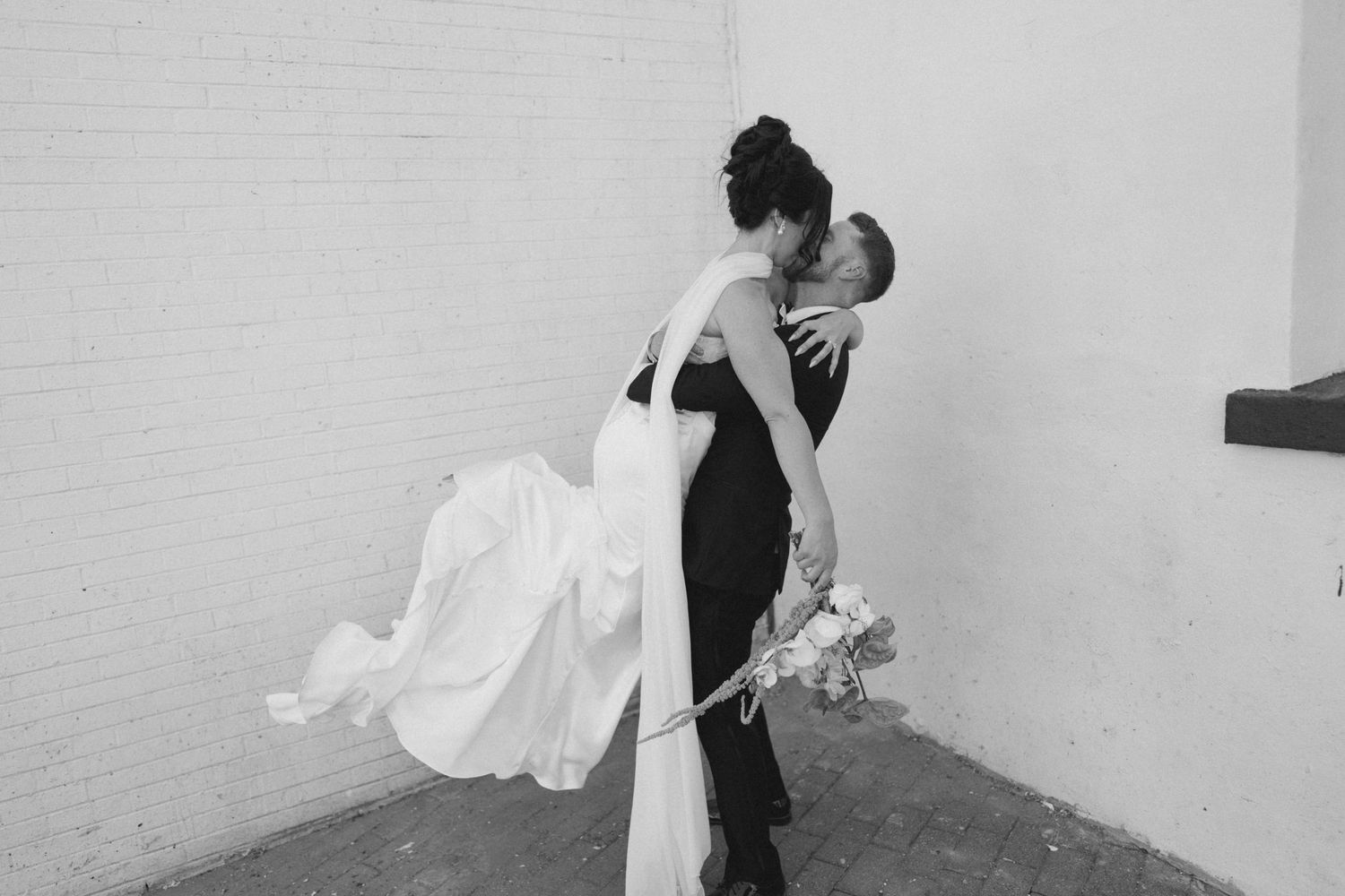 Black and white photo of groom lifting bride in wedding dress, sharing a romantic kiss against a brick wall.