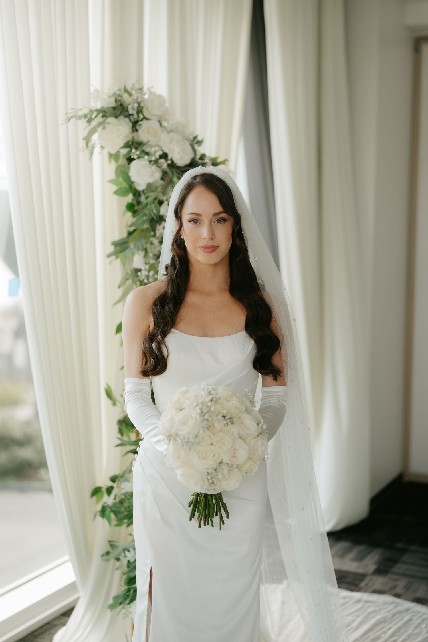 Elegant bride in white satin gown, long gloves, and veil holds white floral bouquet beside a greenery arch and draping curtains.