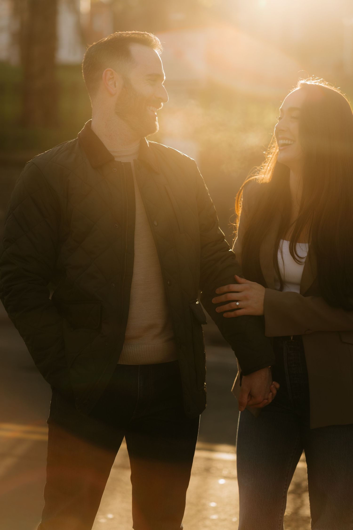 Couple laughing together during golden hour sunset, woman touching man's arm, both wearing dark jackets outdoors.
