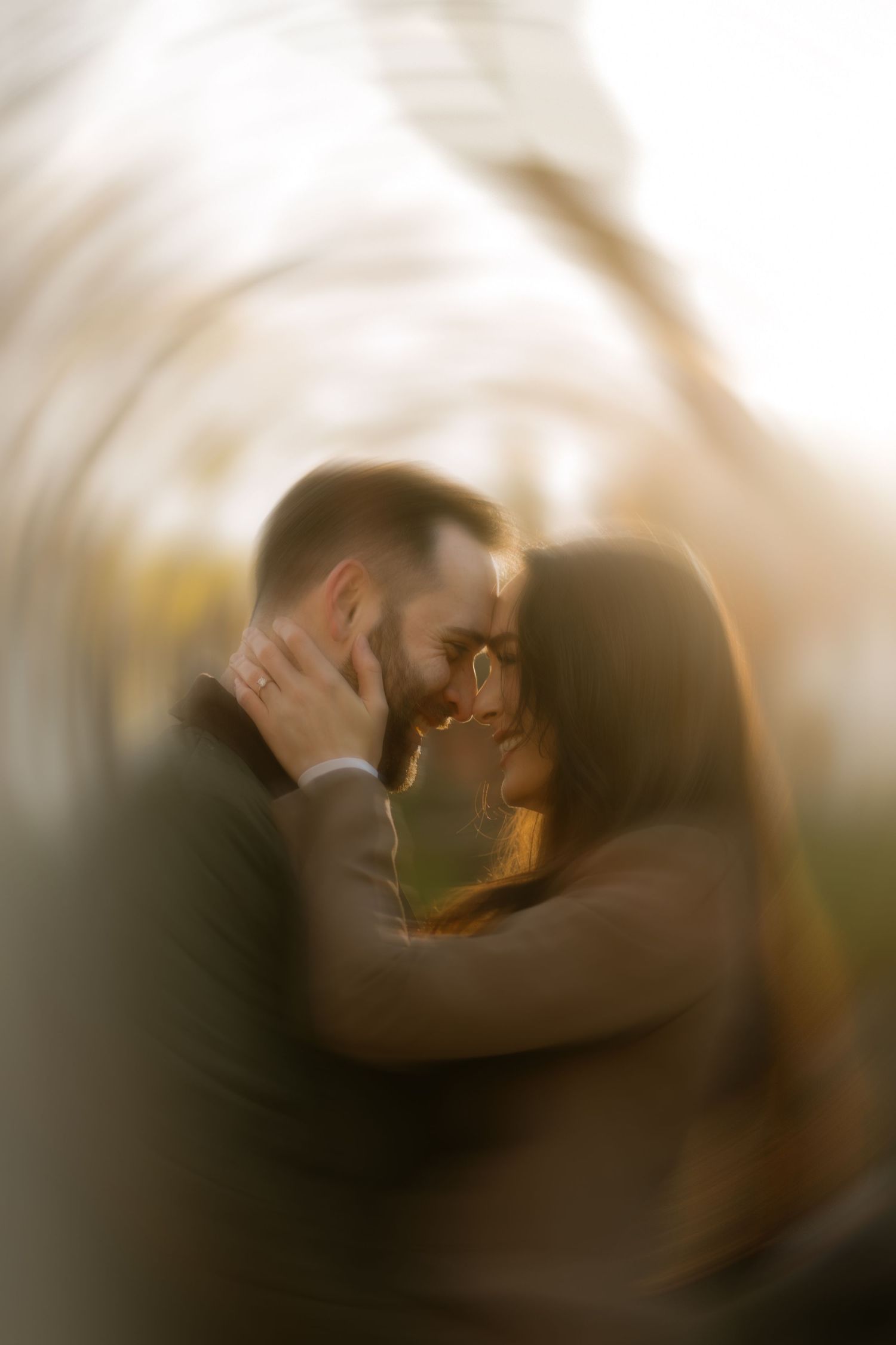 Romantic couple embracing outdoors with dreamy bokeh blur effect during golden hour engagement photo session.